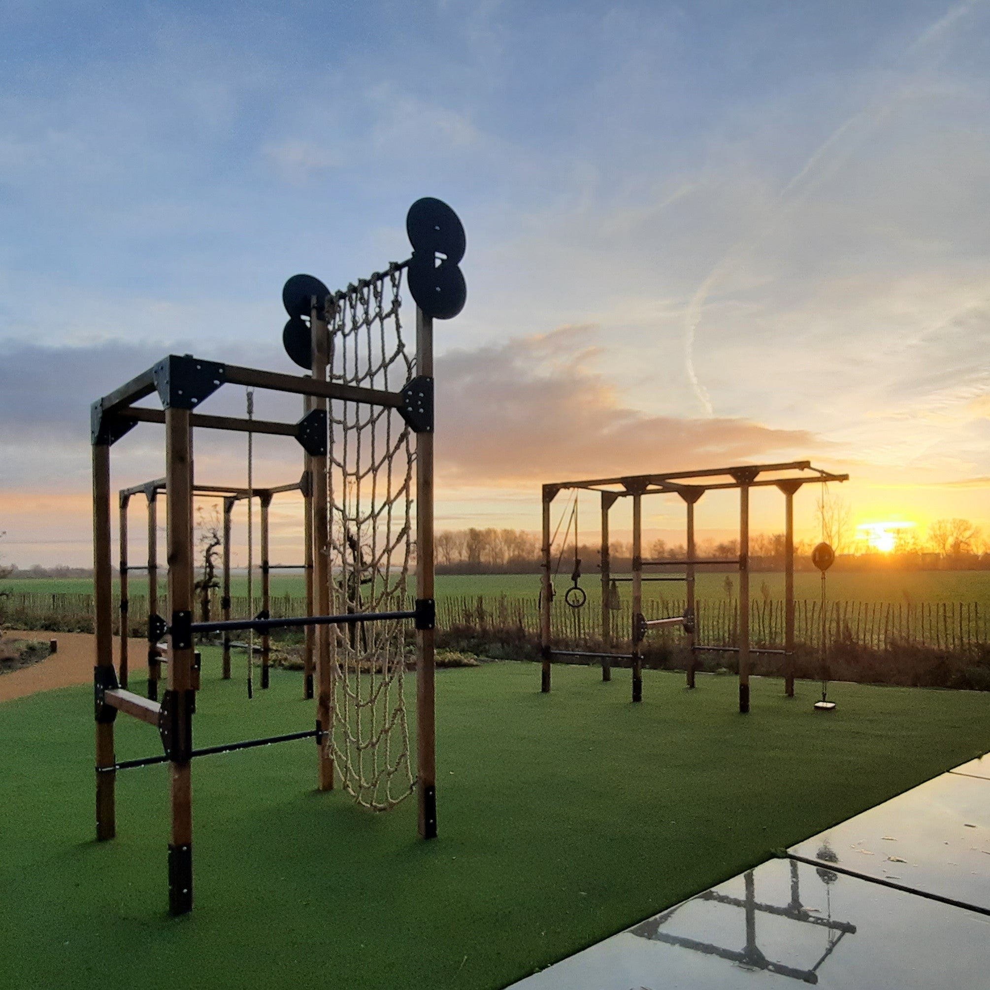 Outdoor fitnesstoestellen, ontworpen voor zowel privé als commercieel gebruik, geplaatst in een groene, open omgeving. De toestellen bestaan uit stevige rekken en stangen, perfect voor verschillende krachttrainingsoefeningen zoals pull-ups, squats, muscle-ups en wall-ball-shots. Gemaakt van duurzaam hout, deze racks bieden een natuurlijke esthetiek die opgaat in de buitenomgeving